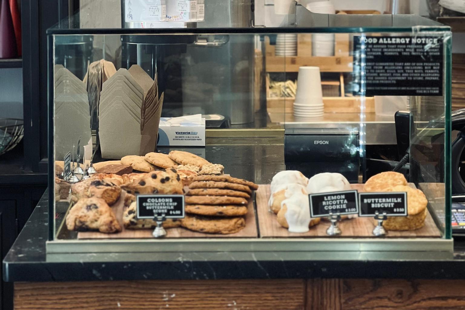 A glass bakery display in New York holds labeled cookies, scones, and biscuits beneath an English food-allergy notice.