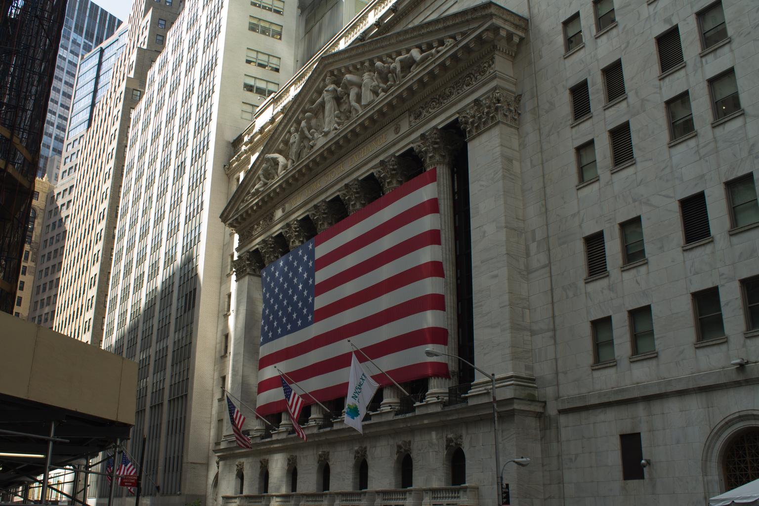 The New York Stock Exchange facade on Wall Street displays a large American flag beneath its carved name.