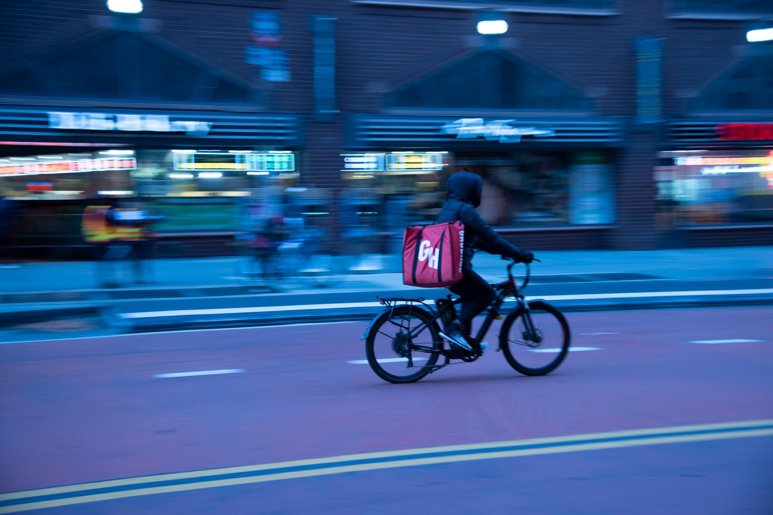 A bicycle delivery courier with a red backpack rides along a New York City street as storefronts blur behind him.