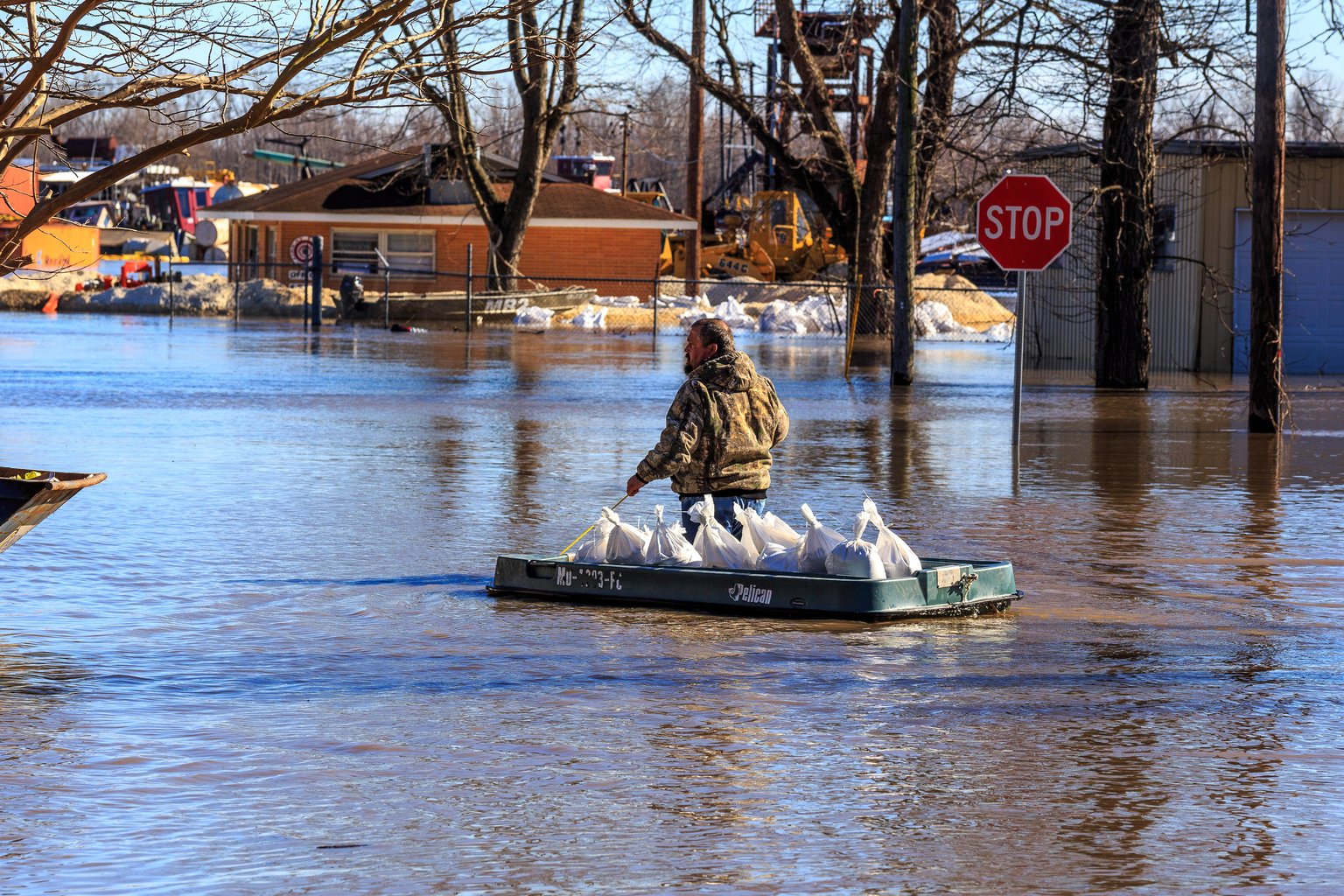 A man pulls a boat loaded with sandbags through floodwater in Cape Girardeau, Missouri, past buildings and a stop sign.